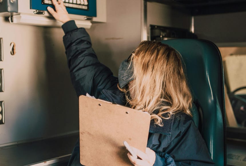 A paramedic in an ambulance adjusts the control panel while holding a clipboard.